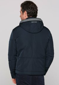 Navy puffer jacket with a hood, featuring a striped collar, quilted shoulder details, and a back logo. Smooth material with a fitted shape.