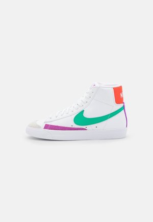 Hvid high-top Nike sneaker med grønt swoosh, lilla og beige accenter og orange hælflap med hvid Nike-tekst på en ensfarvet baggrund.