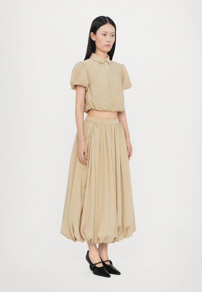 Beige cropped Bluse mit Puffärmeln und Knopfschluss, kombiniert mit einem langen, gerafften Rock aus passendem Stoff und schwarzen spitzen Schuhen.