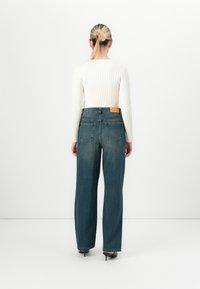 Mulher com cabelo loiro apanhado em um coque, vestindo um suéter branco com texturas, jeans azuis de cintura alta e sapatos de salto pretos, de pé contra um fundo branco liso.