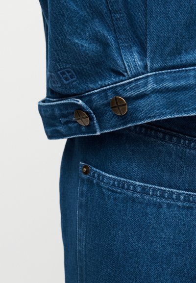 Κοντινό πλάνο μανσετών μπλε τζιν μπουφάν με δύο μεταλλικά κουμπιά και τσέπη μπλε τζιν με καρφί σε απλό φόντο.