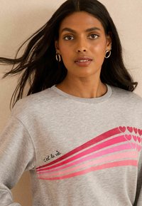 Sudadera gris con gráfico de corazón rosa y franjas. Texto "C'est la vie" bordado. Textura suave, cuello redondo y corte holgado.