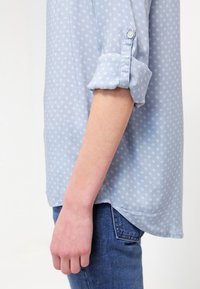 Camicia azzurro chiaro con motivo floreale bianco, maniche arrotolate, dettaglio con bottoni, orlo dritto. Abbinata a jeans blu, che mostrano uno stile informale.