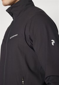 Czarna kurtka softshell z wysokim kołnierzem, zapinana na zamek z przodu i posiadająca kieszeń, z srebrnym logo na piersi oraz odblaskowym akcentem na rękawie.