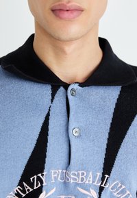 Light blue knitted polo shirt with black accents and two buttons; features cream embroidery of "Fantazy Fussball Club" on the chest.