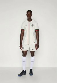 Mees jalgpallur seisab, seljas valge Chelsea FC komplekt, mustad Nike spordijalatsid ja valged sokid, tähistades tasast tausta.