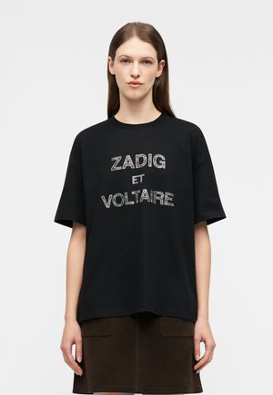 Una giovane donna con lunghi capelli castani indossa una T-shirt nera oversize con la scritta "ZADIG ET VOLTAIRE" e una gonna marrone scuro.