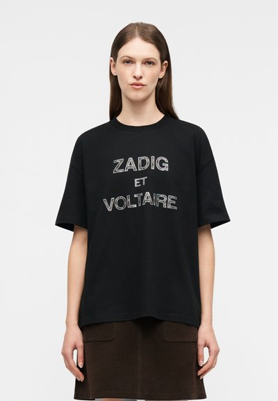 Ung kvinne med langt brunt hår har på seg en svart oversized T-skjorte med teksten "ZADIG ET VOLTAIRE" og et mørkebrunt skjørt.