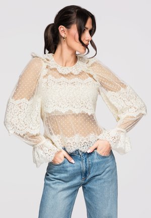 Kobieta w przejrzystej białej koronkowej bluzce w kropki z bufkami, w niebieskich dżinsach, z rękami w kieszeniach, patrząca na bok.