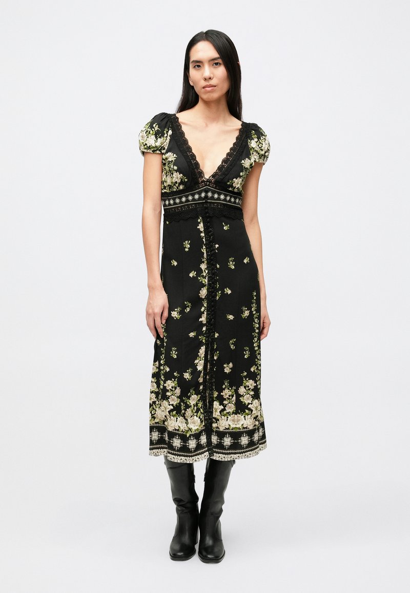 Zwarte bloemenjurk met pofmouwen, diepe V-hals, kantaccenten en borduursel langs de zoom; gecombineerd met zwarte laarzen tot aan de knie.