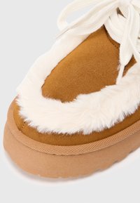 Brune ruskinds sneakers med hvidt faux fur-bemærkning, beige gummisål og cremefarvede snørebånd. Har en afrundet form og teksturerede detaljer.