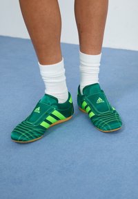 Grønne syntetiske sportsko med et tekstureret design, der har tre hvide striber, polstret pløs og en gummisål. Båret med hvide sokker.