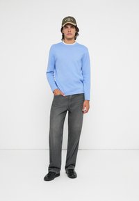 Suéter de malha azul claro com decote redondo, emparelhado com jeans cinza de perna reta e sapatos pretos. Completado com um boné verde oliva.