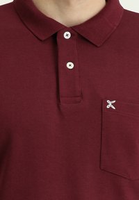 Polo di colore bordeaux realizzato in misto cotone; presenta tre bottoni, una tasca con cuciture decorative e un design del colletto classico.