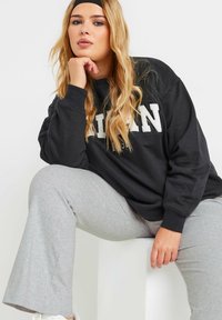 Svart sweatshirt med vit text, avslappnad passform, sänkt axlar; ihopparad med ljusgrå vida byxor och vita sneakers.
