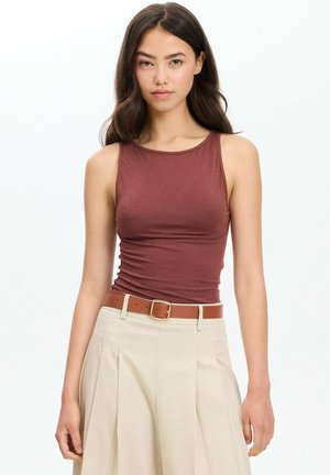 Mujer con cabello largo y oscuro que lleva una blusa marrón sin mangas metida en una falda beige con un cinturón marrón, sobre un fondo liso.