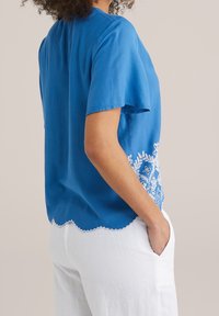 Blå kortärmad blus i lätt material, med vita broderade blommönster längs fållen och klokantade kanter.