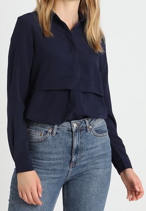 Donna che indossa una camicia blu navy a maniche lunghe abbottonata infilata in jeans blu a vita alta con tasche frontali visibili.