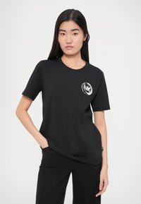 Fekete pamut póló kerek nyakkivágással, rövid ujjakkal és egy fehér kör alakú logóval a bal mellrészen. A modell zsebre tett kézzel pozícionál.