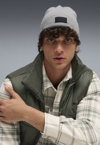 Gray knit beanie with a black logo patch, green puffy vest, and white plaid shirt with gray accents. Close-up of shoulder and face.