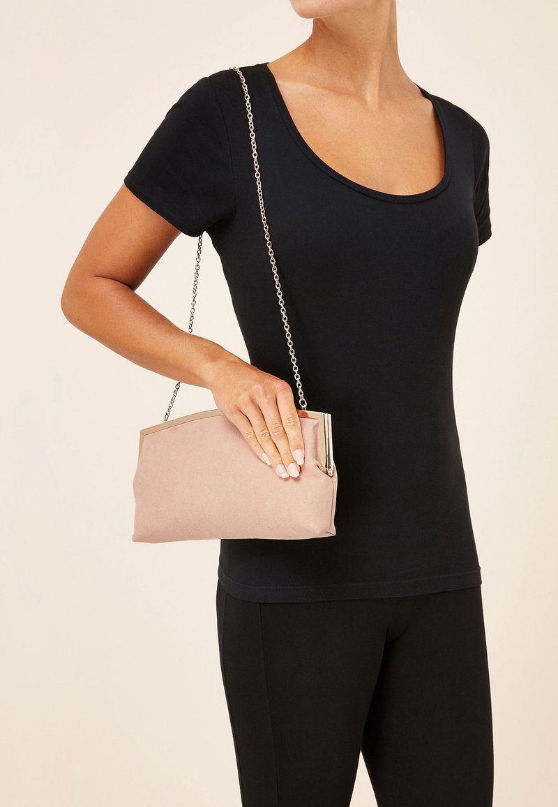Femme portant une tenue noire tenant une petite pochette rose clair avec une bandoulière chaîne argentée sur un fond neutre.