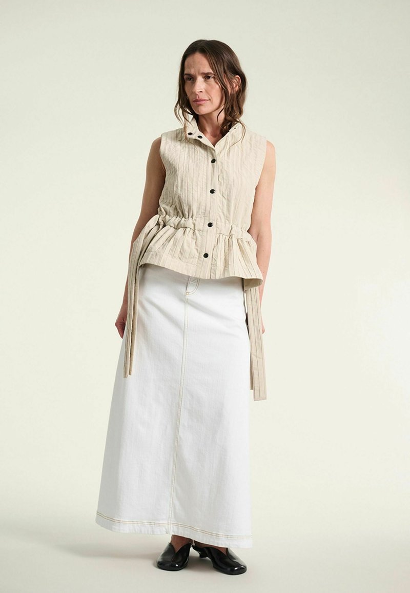Femme portant un haut beige sans manches boutonné avec des liens à la taille, une longue jupe blanche et des chaussures noires, debout devant un fond uni.