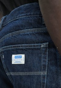 Denimjeans i mørk blå med en teksturert finish, med en hvit merkeetikett. Fremhevet med hvite sømmer og detalj på baklommen.