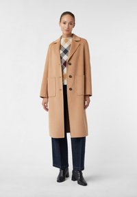 Frau in einem camel-farbenen langen Mantel, kariertem beigen und schwarzen Pullover, dunkelblauen weiten Jeans und schwarzen Stiefeletten vor weißem Hintergrund.