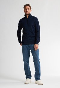 Navy zip-nekwol gemaakt van zachte stof, met lange mouwen en geribbelde manchetten, gecombineerd met lichtblauwe rechte jeans en witte sneakers.