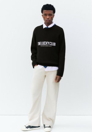 Jeune homme portant un pull noir avec le texte "THE LUCKY CLUB", une chemise blanche, un pantalon large beige et des baskets noires et blanches, debout contre un mur blanc.