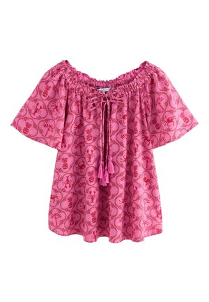 Eine Frau trägt eine rosa gemusterte Bluse mit kurzen Ärmeln und Jeansshorts und steht an einer strukturierten Steinmauer.
