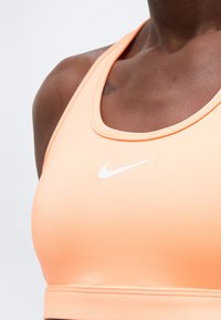 Κοντινό πλάνο ενός ατόμου που φοράει ροδάκινο αθλητικό σουτιέν Nike με λευκό λογότυπο στο στήθος, σε φωτεινό φόντο.