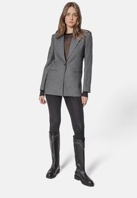 Blazer texturat gri cu un singur nasture, purtat peste un top transparent negru. Leggings negri ajustați și cizme negre până la genunchi cu accente decorative.
