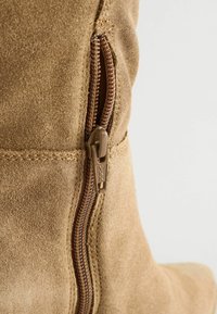 Tan suede saapas, jossa on sivuvetoketju ruskeilla yksityiskohdilla, ommeltuja yksityiskohtia ja teksturoitu pinta. Muotoilu on vartalonmyötäinen ja tyylikäs.