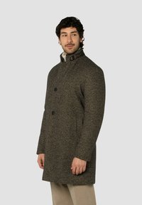 Brauner Tweedmantel mit hohem Kragen, ausgestattet mit einer Knopfleiste vorne, Seitentaschen und einem strukturierten Stoff. Akzentuiert mit einem dunklen Kragenriemen.