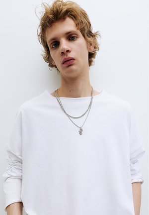 Jong persoon met lichtbruin haar, een losse witte lange mouw shirt en twee zilveren kettingen tegen een effen achtergrond.