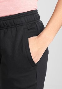 Černé joggingové kalhoty s elastickým pasem, které mají boční kapsy. Materiál se zdá být hladký a prezentuje jednoduchý design bez vzorů.