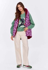 Kobieta w lśniących beżowych spodniach, zielonej bluzie z graficznym motywem czarnego psa, fioletowej kamizelce w kwiaty i zielono-fioletowych sneakersach, stoi z jedną ręką w kieszeni.