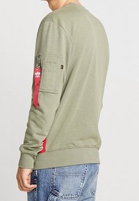 Sudadera verde oliva con cuello redondo, bolsillo lateral, tirador de cremallera rojo y puños de canalé. Presenta un ajuste relajado y una textura suave.