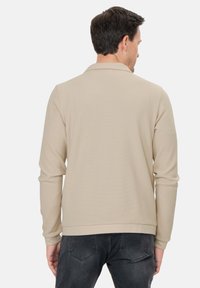 Beige strukturiertes Langarmhemd mit Kragen und gerippten Manschetten, kombiniert mit dunklen Jeans. Auffälliges Netzstoffmuster. Ansicht von hinten.