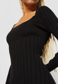 Robe noire côtelée à manches longues avec un décolleté carré et une coupe ajustée. Comprend un petit logo brodé sur le côté. Tissu texturé.