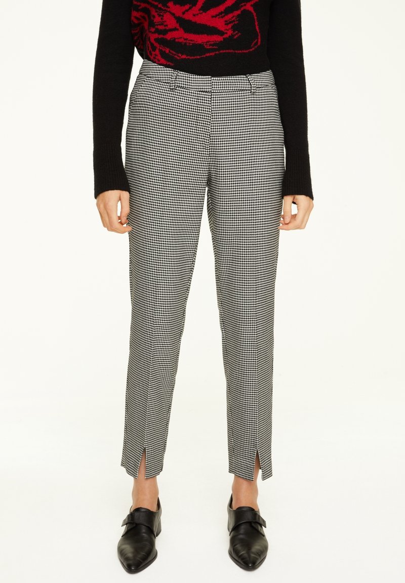 Femme portant un pantalon cheville pied-de-poule noir et blanc avec des fentes à l'avant, un pull noir avec un motif rouge, et des chaussures noires pointues.