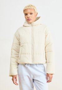 Casaco puffer cropped bege com capuz, apresentando um fecho de correr frontal, bainha elástica e costuras visíveis. Tecido leve com uma textura suave.