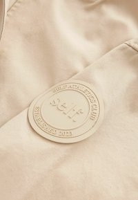 Il tessuto beige presenta un logo circolare in rilievo beige con la scritta "self." e "SELF ATHLETICS CLUB ESTABLISHED 2023" impresso.