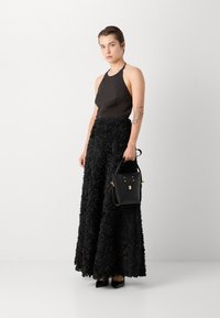 Haut noir à dos nu avec une jupe maxi noire à fleurs texturées. Tient un petit sac à main noir avec des accents dorés et des talons noirs à bout pointu.