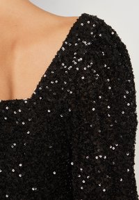 Svart paljett-topp med genomskinligt tyg, med fyrkantig halsringning och puffärmar. Tätt packade svarta paljetter täcker ytan och reflekterar ljus.