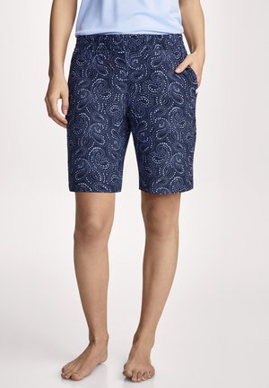 Vrouw draagt marineblauwe korte broek met witte stippen in een spiraalvormig patroon, staat op blote voeten met één hand in de zak tegen een effen achtergrond.