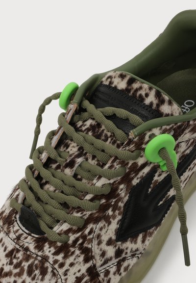 Gros plan d'une basket avec un motif tacheté marron et blanc, des lacets vert olive, des bloque-lacets verts et des détails latéraux noirs.