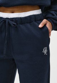 Mano in tasca, indossa pantaloni sportivi blu navy con cordoncino bianco, elastico bianco in vita e logo ricamato sulla coscia.