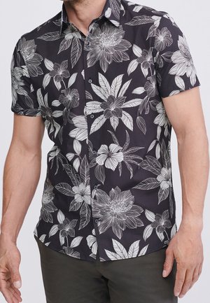 Chemise noire à manches courtes boutonnée avec des motifs floraux et feuillages blancs détaillés, portée par une personne portant un pantalon vert olive.